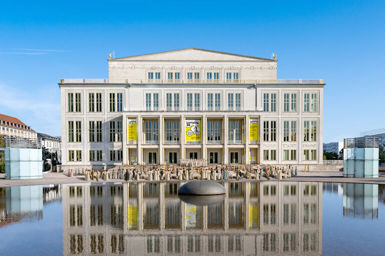 Hinter dem Brunnen auf dem Augustusplatz sieht man das Opernhaus von Leipzig und viele Menschen in historischen Kostümen.