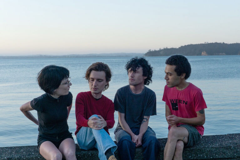 Das Quartett sitzt auf einer Mauer, hinter sich das Meer und einen Küstenstreifen. Die Köpfe der Musikerin und der Musiker wurden grafisch vergrößert.