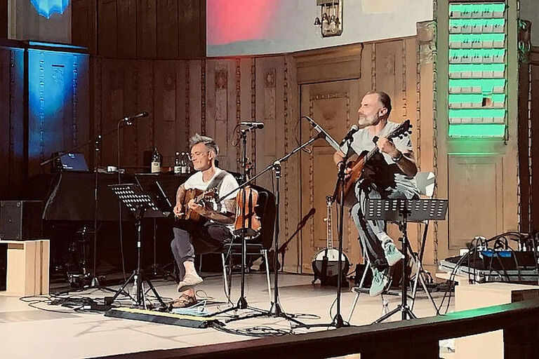 Zwei Musiker sitzen in einem holzvertäfelten Raum, spielen Gitarre und singen.