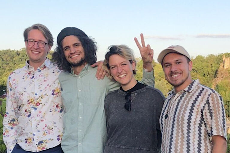 Gruppenfoto der lachenden Bandmitglieder an einem Aussichtspunkt mit Felsen und Wald im Hintergrund