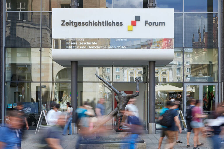 Außenansicht des Museums mit seiner spiegelnden Glasfassade und vielen Passanten sowie einer Skulptur davor