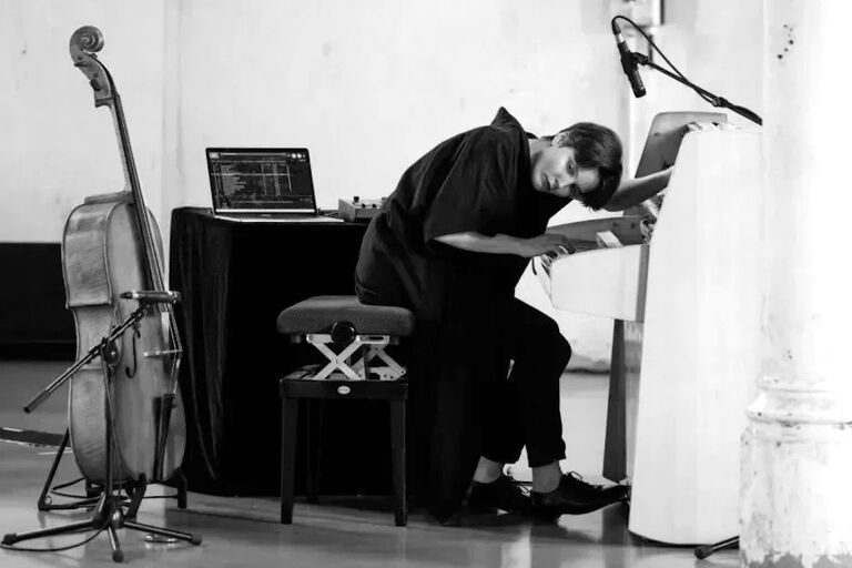 Die Künstlerin spielt weit nach vorn gebeugt an einem Tasteninstrument, hinter sich einen Laptop und ein Cello
