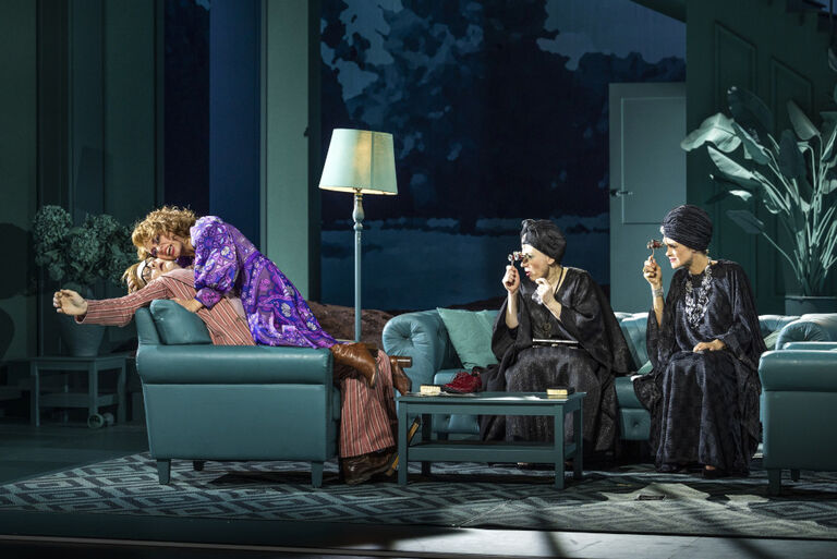 Zwei ältere Damen sitzen auf einem Sofa und schauen lachend interessiert durch ihre Operngläser zu, wie eine Frau eine leblose Person umarmt.