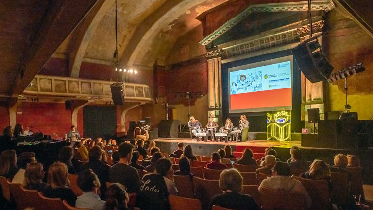 Foto einer Pressekonferenz zur Filmwoche im UT Connewitz, einem historischen Kino. Die Veranstalter sitzen an einem Tisch auf der Bühne und die Journalisten sind im Zuschauerraum.