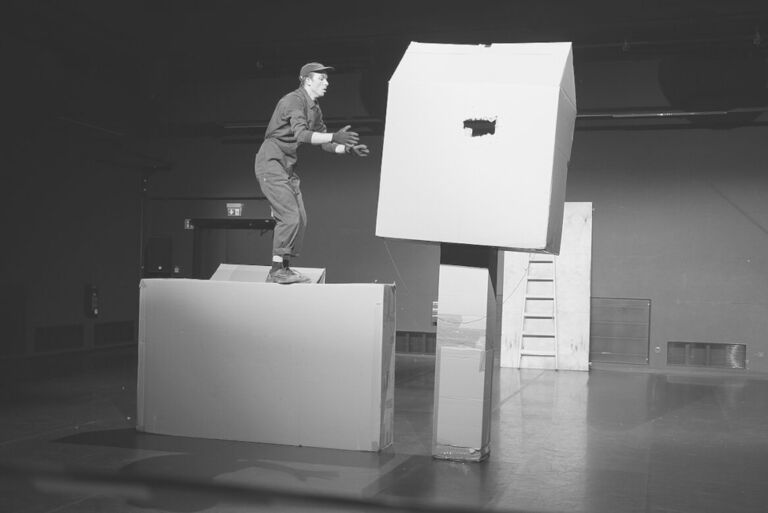 Foto des Schauspielers der in Arbeitskleidung mit großen Kisten aus Pappe hantiert.