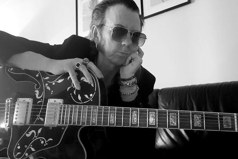 Der Musiker sitzt mit seiner verzierten Gitarre auf einem Sofa, hat das Kinn auf eine Hand gestützt und schaut nachdenklich vor sich hin.