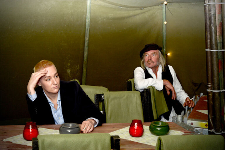 Katharina Pongratz und Jens-Paul Wollenberg sitzen an verschiedenen Tischen mit Windlichtern und Aschenbechern, rauchen und schauen teilnahmslos in die Luft.