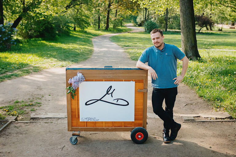 Der Musiker steht mit seinem Klavier auf Rollen in einem grnen Park und lächelt in die Kamera.