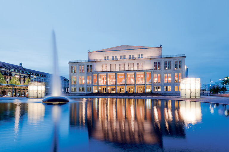 Das festlich erleuchtete Gebäude der Oper mit Wasserbecken und Fontaine davor in der Dämmerung
