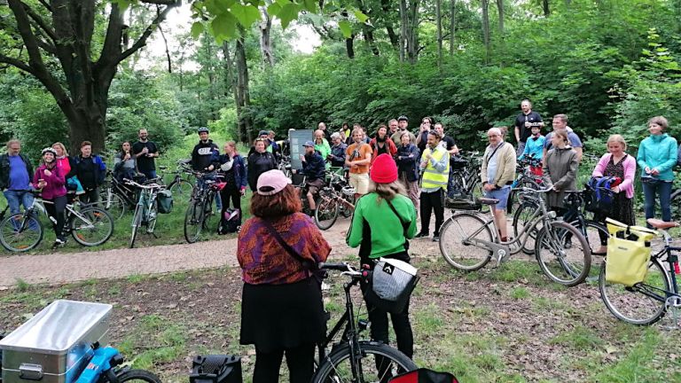 Auf einem Waldweg stehen viele Menschen mit Fahrrädern und hören einem Autor zu.
