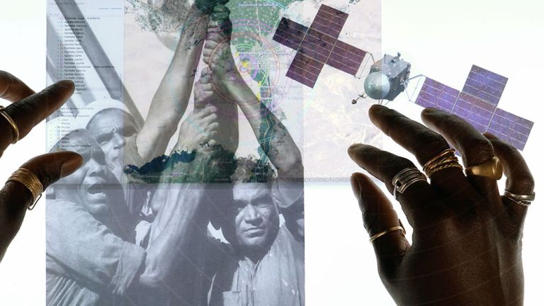 Auf einem Licht-Tisch liegen verschiedene Fotos übereinander, das einer Karte, von drei Männrn und das eines Satelliten