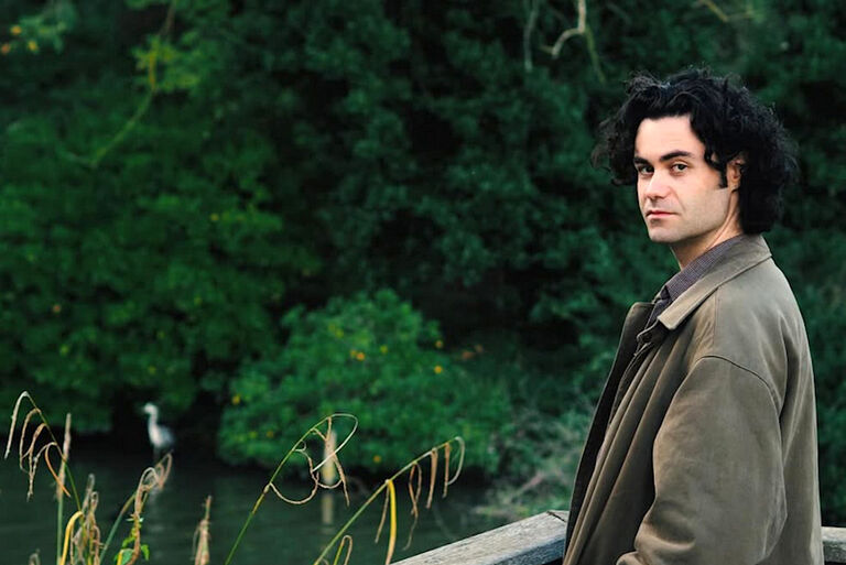 Der Musiker steht mit nachdenklichem Blick an einem Holgeländer vor einem Teich.