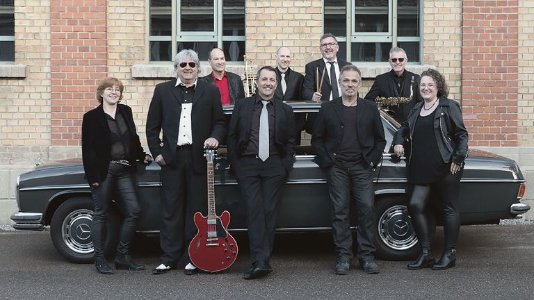 Gruppenfoto der Musikerinnen und Musiker, die in schwarzer Kleidung um einen alten Mecedes herumstehen