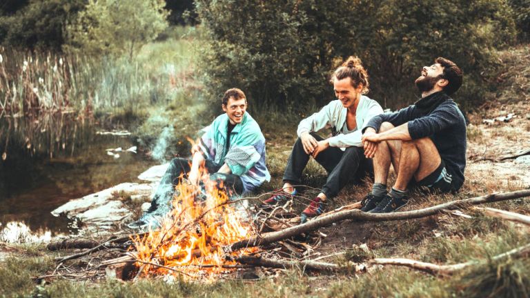 Drei junge Männer sitzen lachend um ein Lagerfeuer an einem kleinen See.