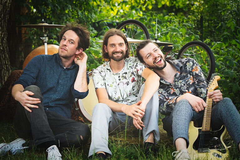 Die drei Musiker sitzen mit ihren Instrumenten auf einer Wiese. Im Hintergrund sind Büsche und Bäume zu sehen.