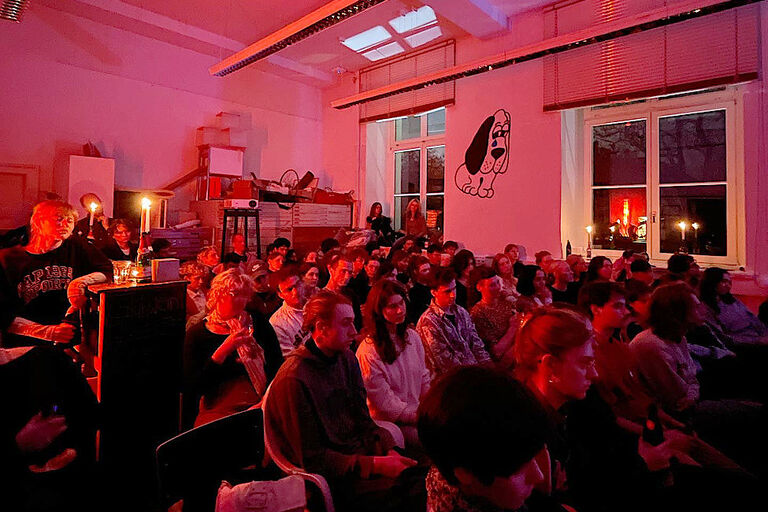 Blick in einen rot beleuchteten Raum mit Tresen und vielen Menschen, die auf Stühlen sitzend eine Lesung verfolgen.