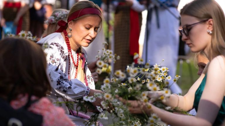 Eine Frau in ukrainischer Tracht bindet mit einer Besucherin an einem Stand einen Blumenkranz.