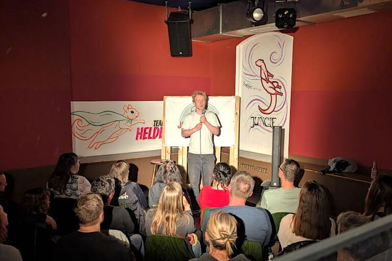 Ein Comedian steht ein einem kleinen Raum mit Mikrofon vor Publikum.