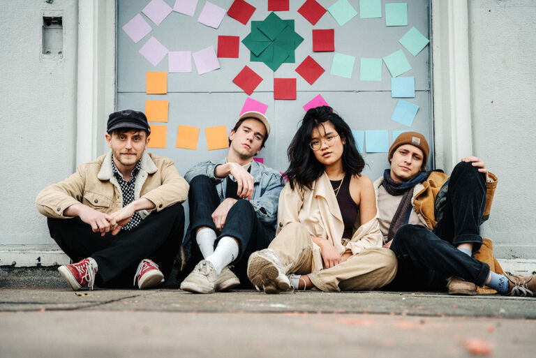 Die Band sitzt auf dem Boden vor einer Wand, die mit bunten Klebezetteln verziert ist.