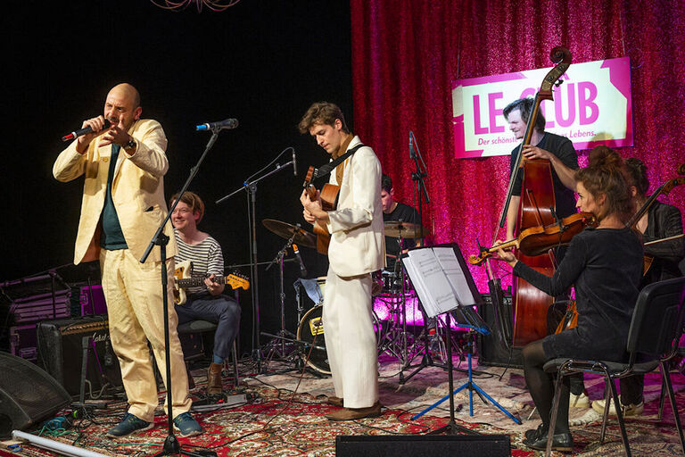 Auf einer Bühne vor einer Band steht ein Moderator in einem gelben Anzug.