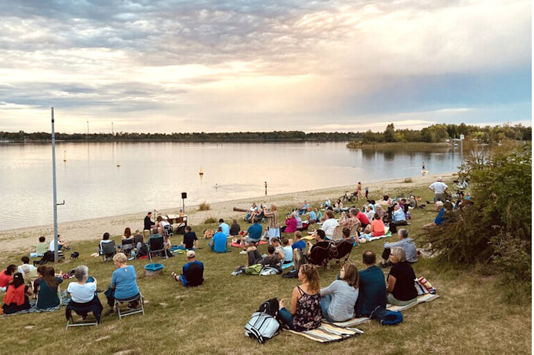 Viele Menschen sitzen auf Decken oder Camping-Stühlen am Ufer eines Sees und lauschen einem Piano-Konzert.