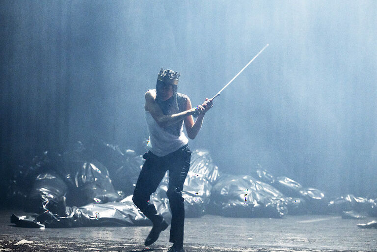 Über eine einer Bühne voller Müllsäcke lauft eine Schauspielerin mit Krone und erhobenem Schwert.