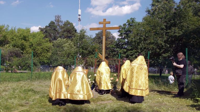 Fünf Geistliche in goldenen Umhängen stehen vor einem Holzkreuz auf einer Wiese, während rundherum die Menschen im Park ihre Freizeit verbringen.