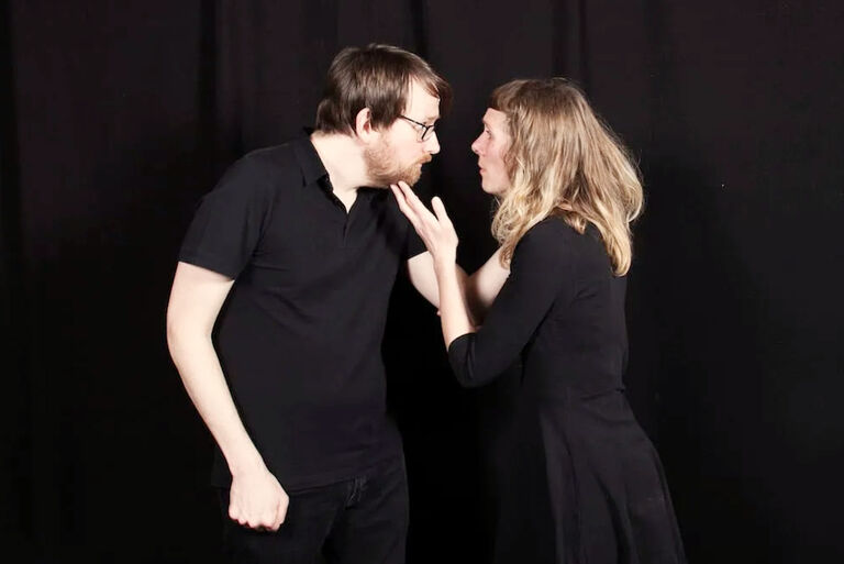 Ein Schauspieler und eine Schauspielerin stehen sich wie ein Liebespaar gegenüber, schauen sich aber eher verwundert an.