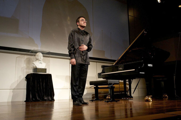 Der Pianist steht nach einem Konzert neben seinem Flügel und bedankt sich mit einer Geste beim Publikum.