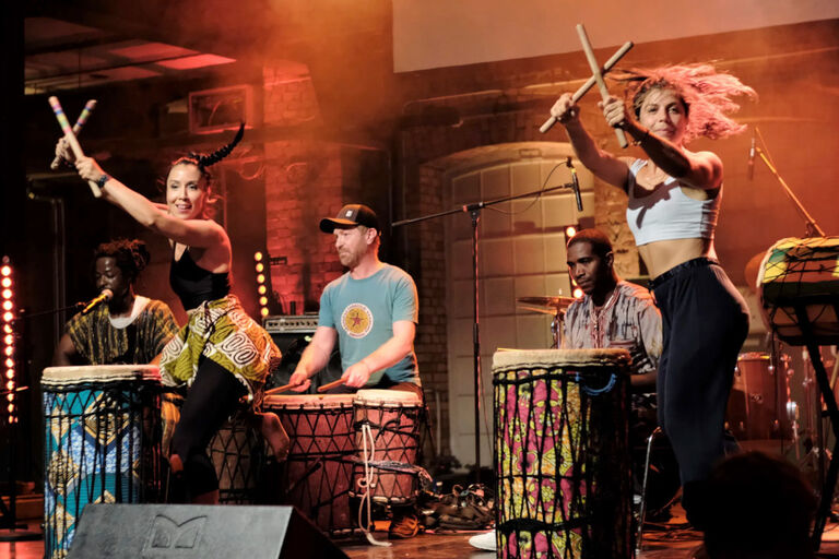 Zwei Tänzerinnen mit klanghölzern tanzen vor einer Gruppe aus drei Percussionisten