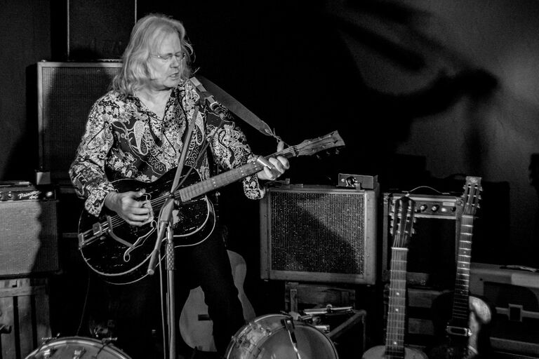 Der Musiker steht bei einem Auftrit mit seiner Gitarre zwischen mehreren Boxen und Instrumenten.