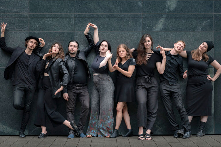 Die jungen Schauspielerinnen und Schauspieler stehen in extravagenten Posen und in Dunkler Kleidung vor einer ebenfalls dunklen Wand.