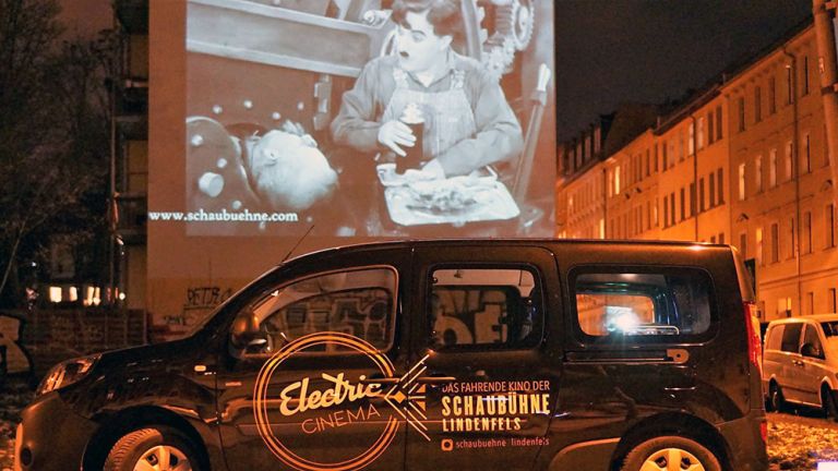 Das Auto "Electric Cinema" steht nachts auf der Straße und projetziert eine Filmszene mit Charlie Chaplin auf eine Giebelwand.