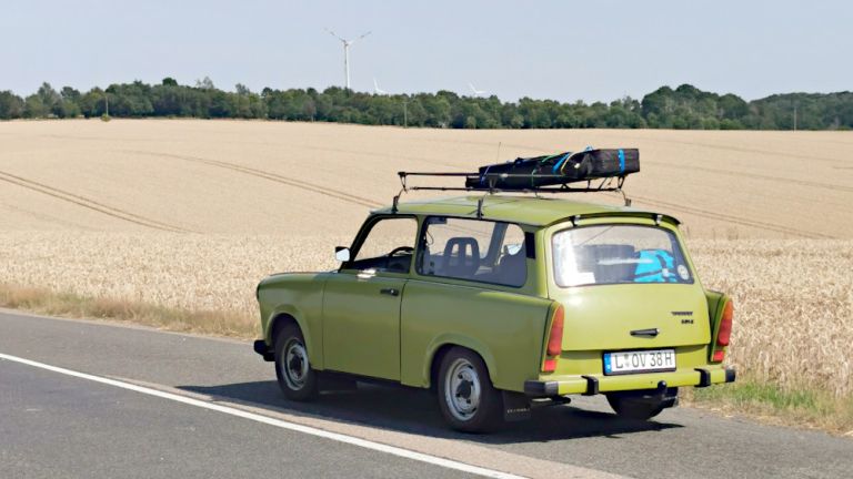 Ein grüner Trabant Kombi fährt auf einer Landstraße an einem Getreidefeld entlang.
