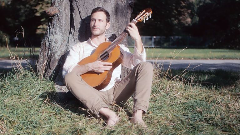 Der Musiker sitzt mit seiner Gitarre an einen Baum gelehnt auf einer sommerlichen Wiese.