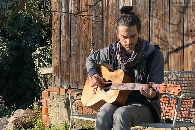 Der Musiker sitzt auf einer Bank vor einem Holzschuppen und spielt Gitarre.