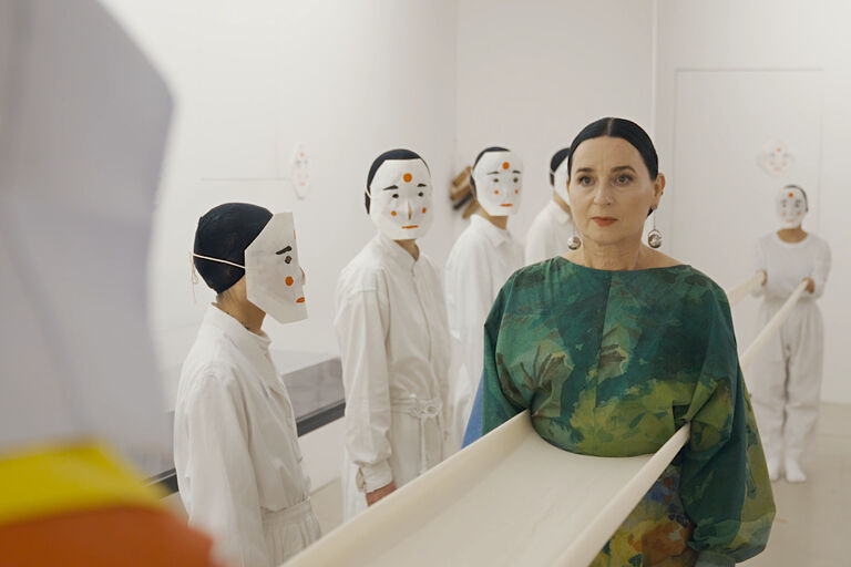 Eine Frau im grünen Kleid steht zwischen weißgekleideten Personen mit weißen Papiermasken vor den Gesichtern.
