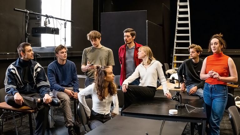 Acht Studierende sitzen und stehen in einem Proberaum beieinander
