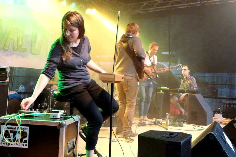 Das Trio bei einem Auftritt auf der Bühne. Im Vordergrund sitzt eine Frau an dem elektronischen Instrument Theremin.