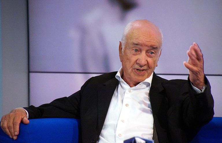 Armin Müller-Stahl sitzt bei einer Veranstaltung auf einem Sofa und spricht, während er eine Hand erhoben hat.
