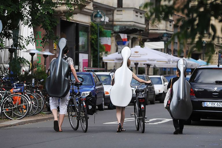 Drei junge Musikerinnen und Musiker laufen mit großen Cellokoffern auf dem Rücken eine Straße entlang.