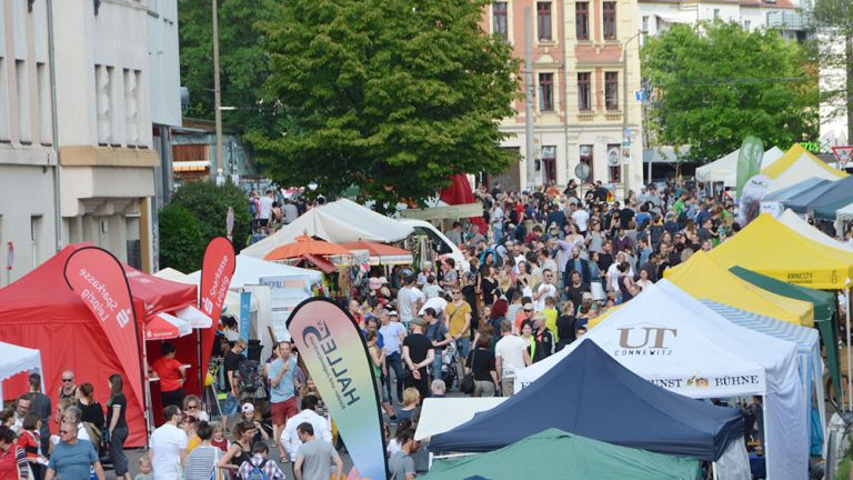 Auf einer Straße im Wohngebiet sind links und rechts Stände und Pavillons aufgebaut, zwischen denen eine große Menschenmenge flaniert.