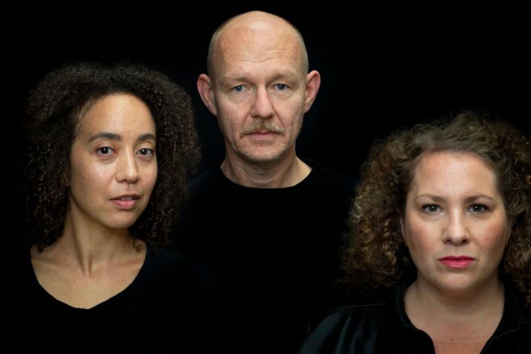 Foto von drei ernst blickenden Ensemblemitgliedern in schwarzer Kleidung und vor schwarzem Hintergrund