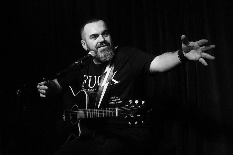 Bei einem Konzert sitz der Sänger mit seiner Gitarre vor einem Mikrofon und streckt seinen Arm aus.