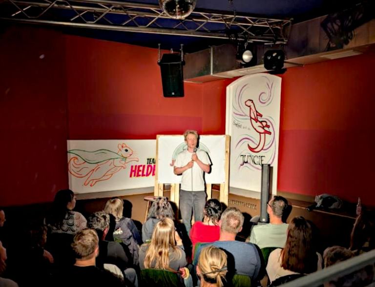 Ein Comedian steht ein einem kleinen Raum mit Mikrofon vor Publikum.