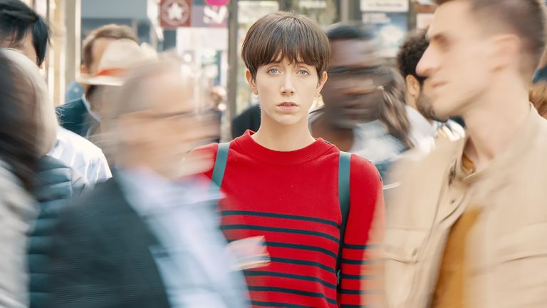 Filmszene, in der die Hauptdarstellerin in rotem Pullover und mit kurzen braunen Haaren auf Straße ganz still zwischen vielen Menschen steht, die alle in Bewegung sind.