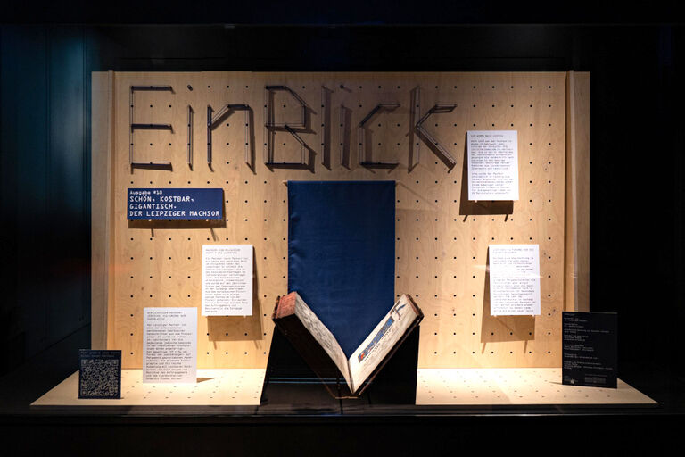 Ein dickes historisches Buch wird in einer Vitrine mit meheren Tafeln mit erläuterungen ausgestellt