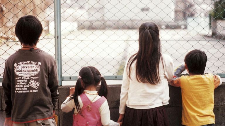 Vier Kinder unterschiedlicher Größe stehen vor einem Drahtzaun und schauen hindurch auf eine kahle Fläche.