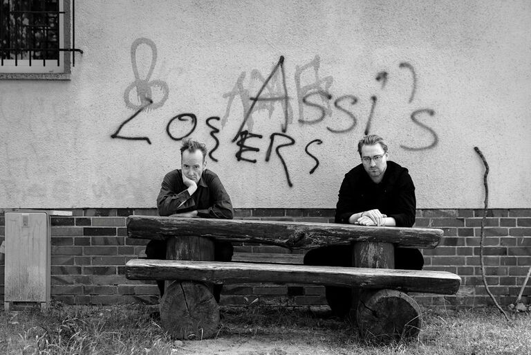 Die beiden Autoren sitzen an einem rustikalen Holztisch vor einer Wand mit den Graffitis "Assi's" und "Losers".