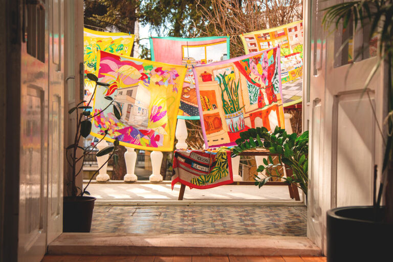 Durch eine geöffnete Flügeltür sieht man farbige Tücher mit vielfältigen Motiven, die auf Wäscheleinen hängen.